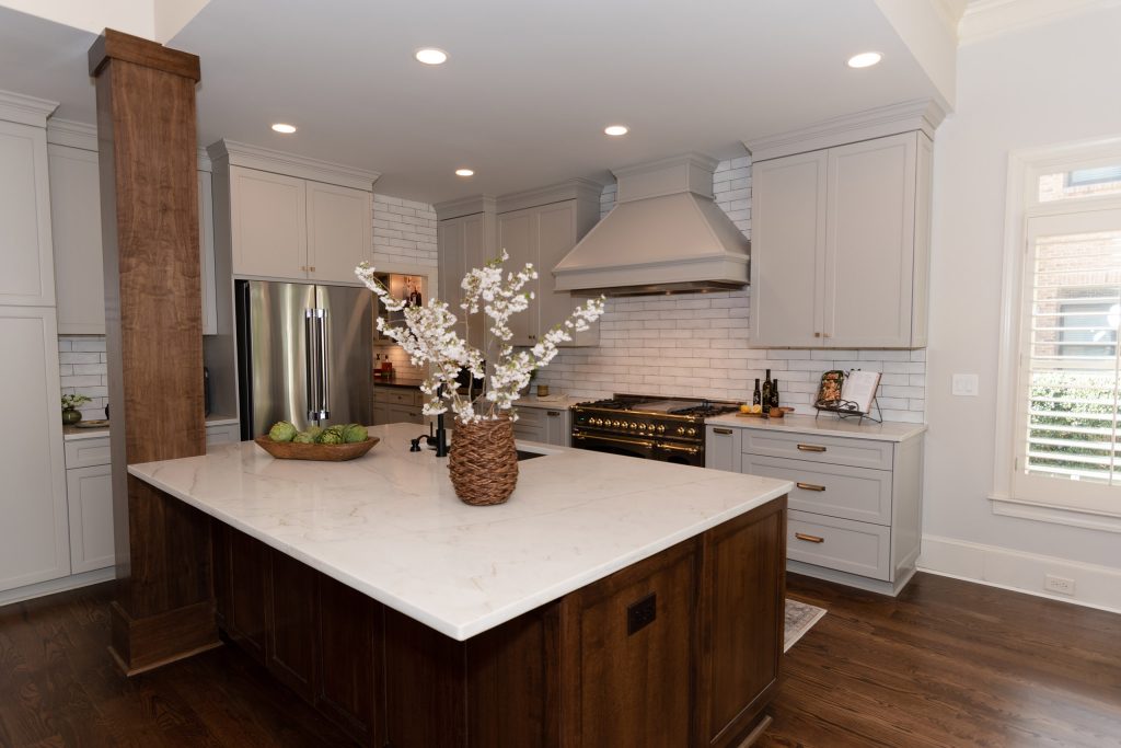 Walnut peninsula island with white quartz top, gray cabinets, painted hood, and a black-and-brass range beyond.