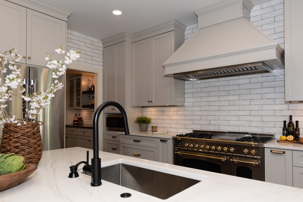White quartz island with a black faucet facing a black-and-brass range, painted hood, and light-gray cabinets.