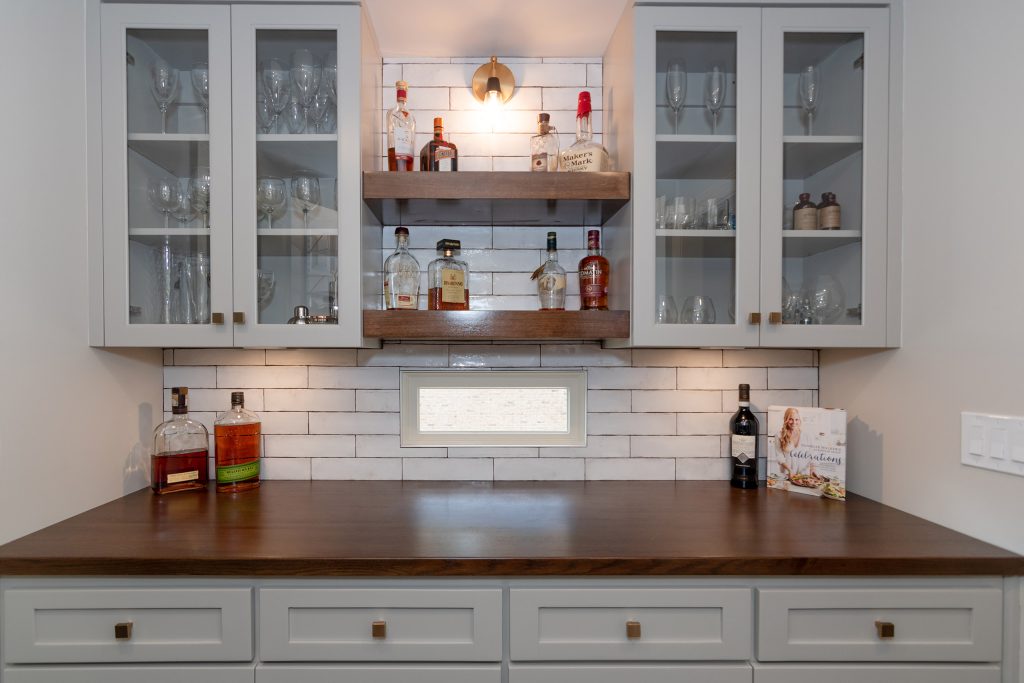 Corner bar with white shaker and glass-front cabinets, floating walnut shelves, and a walnut counter over white subway tile.