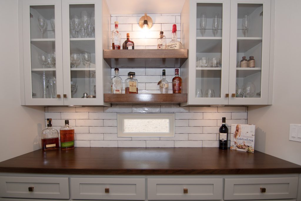 Bar wall with white glass-door cabinets, floating walnut shelves of spirits, walnut counter, and white subway tile.