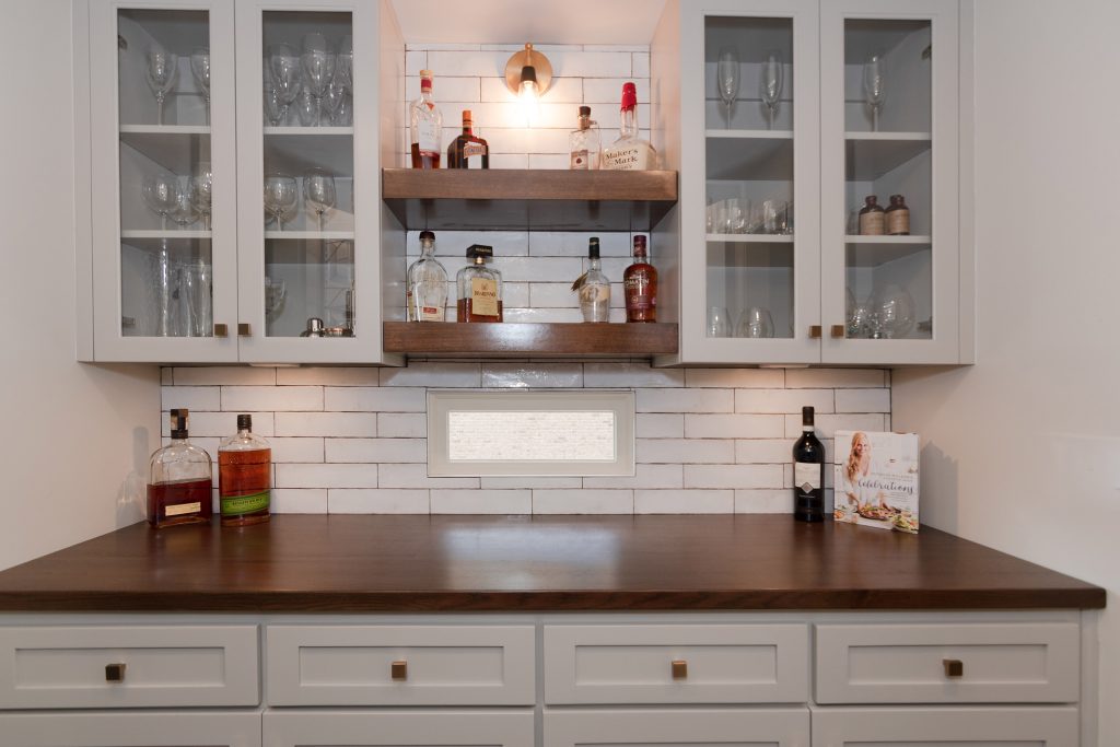Symmetrical bar hutch with glass-front white cabinets, two floating walnut shelves, and a walnut counter under a brass sconce.