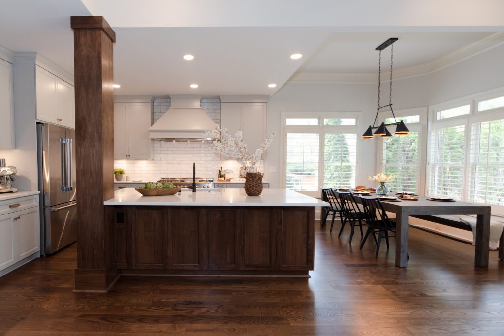 Open kitchen with a walnut island and column, light-gray perimeter cabinets, painted hood, and a black-and-brass chandelier over the table.