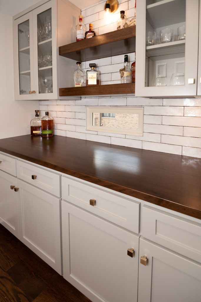 Beverage bar with white glass-front cabinets, floating walnut shelves, walnut counter, and white subway tile.