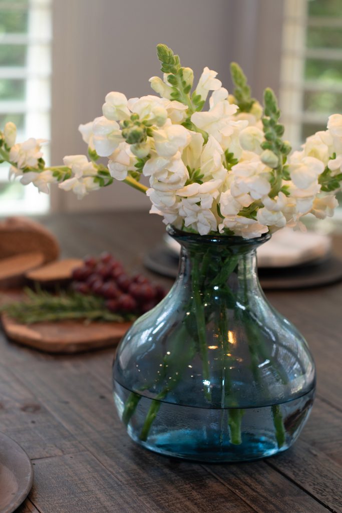 Recycled blue-glass vase of white snapdragons on a dark reclaimed-wood table with grapes and rosemary behind.
