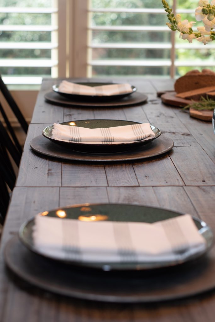 Table place settings with pewter chargers, dark plates, and striped napkins running down a reclaimed-wood table.
