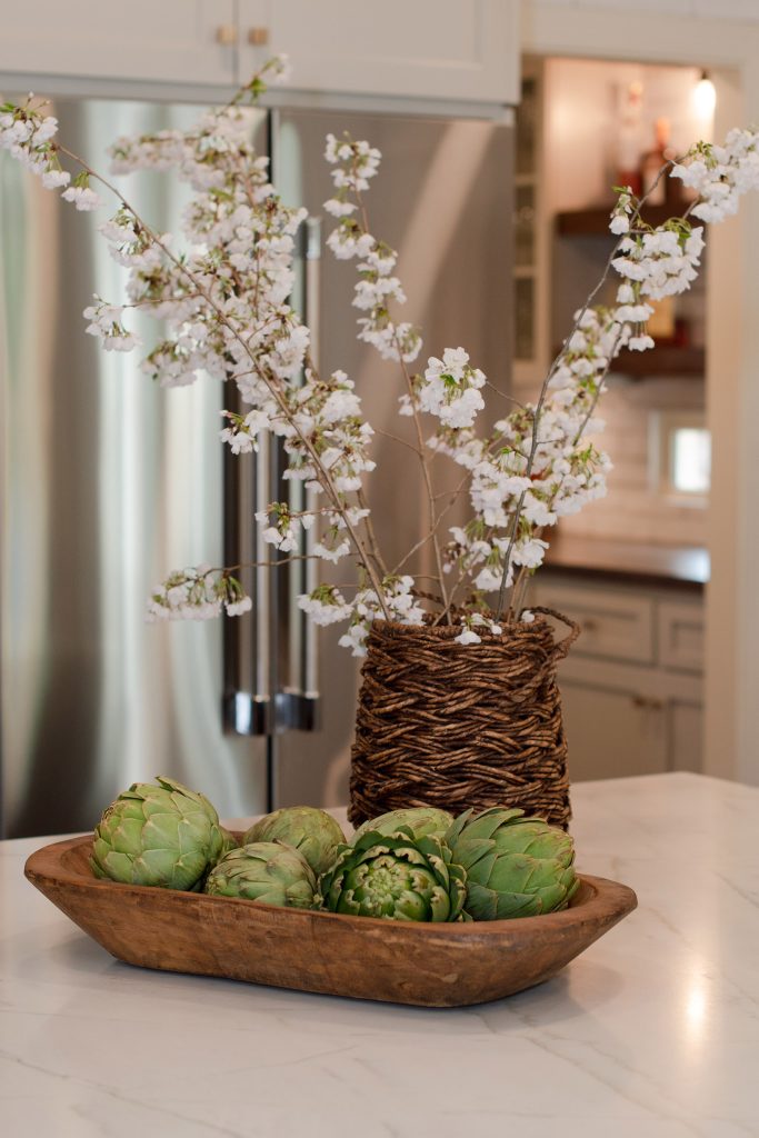 Wood dough bowl of artichokes and a wicker vase of white cherry blossoms on a white quartz counter before a stainless fridge.