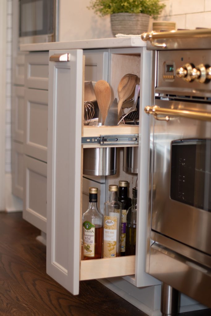 Pull-out utensil and oil cabinet beside a stainless pro range with brass knobs and handle rails.