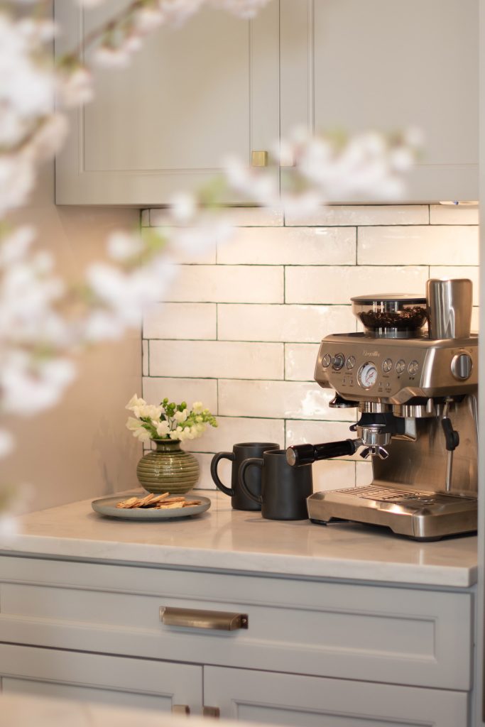 Coffee station with a stainless espresso machine, gray cabinets, white quartz counter, and glazed white subway tile.