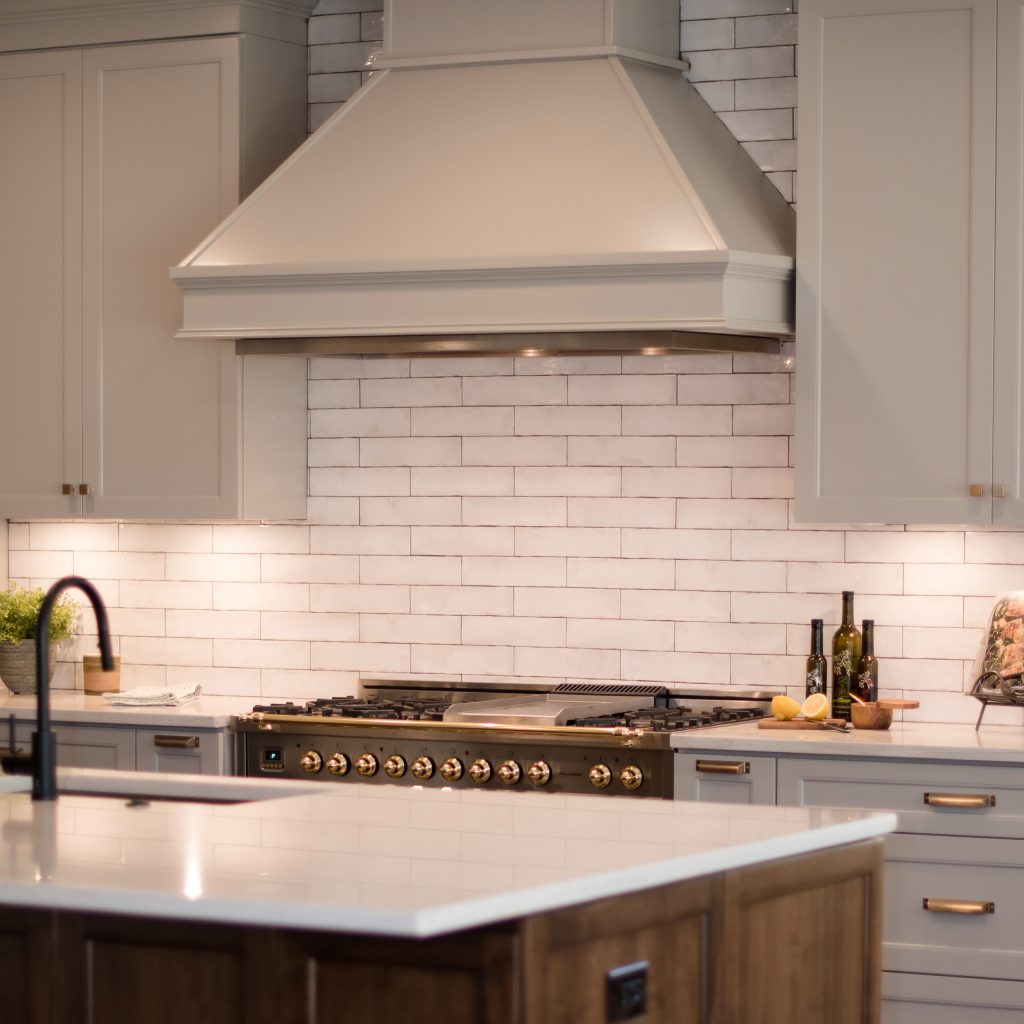 Painted vent hood over a black-and-brass range, framed by handmade white subway tile and gray cabinets.