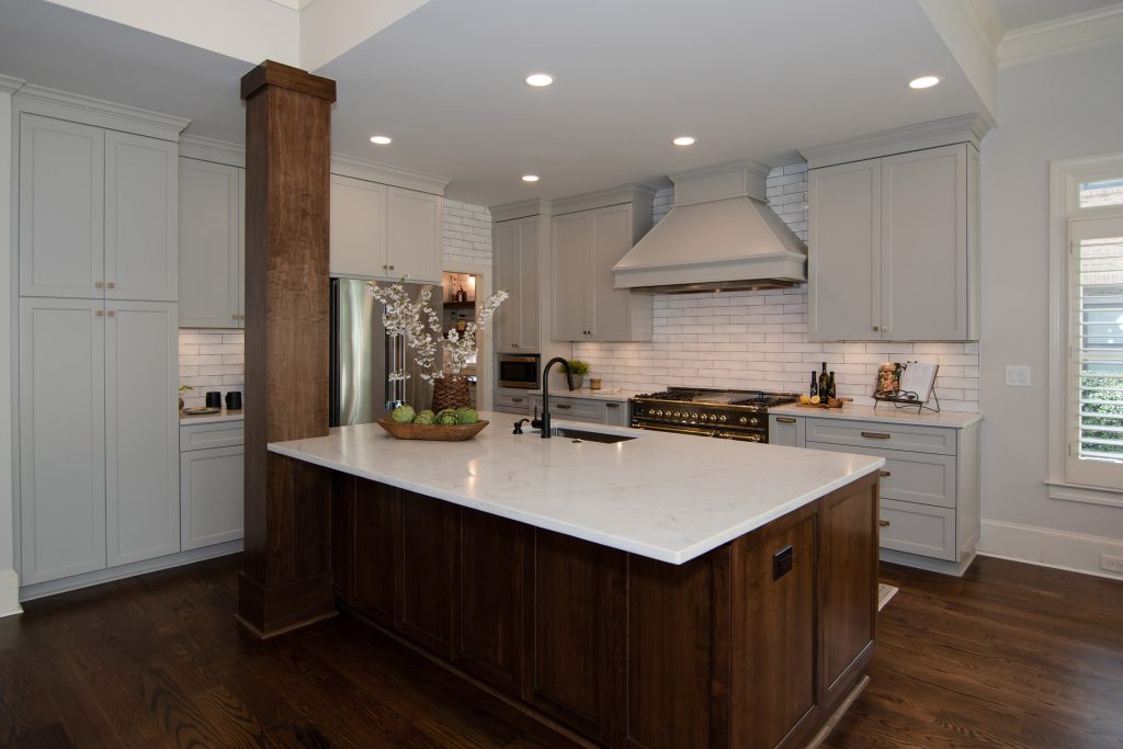 Walnut island and column with white quartz top, painted vent hood, and black-and-brass range against light-gray cabinets.