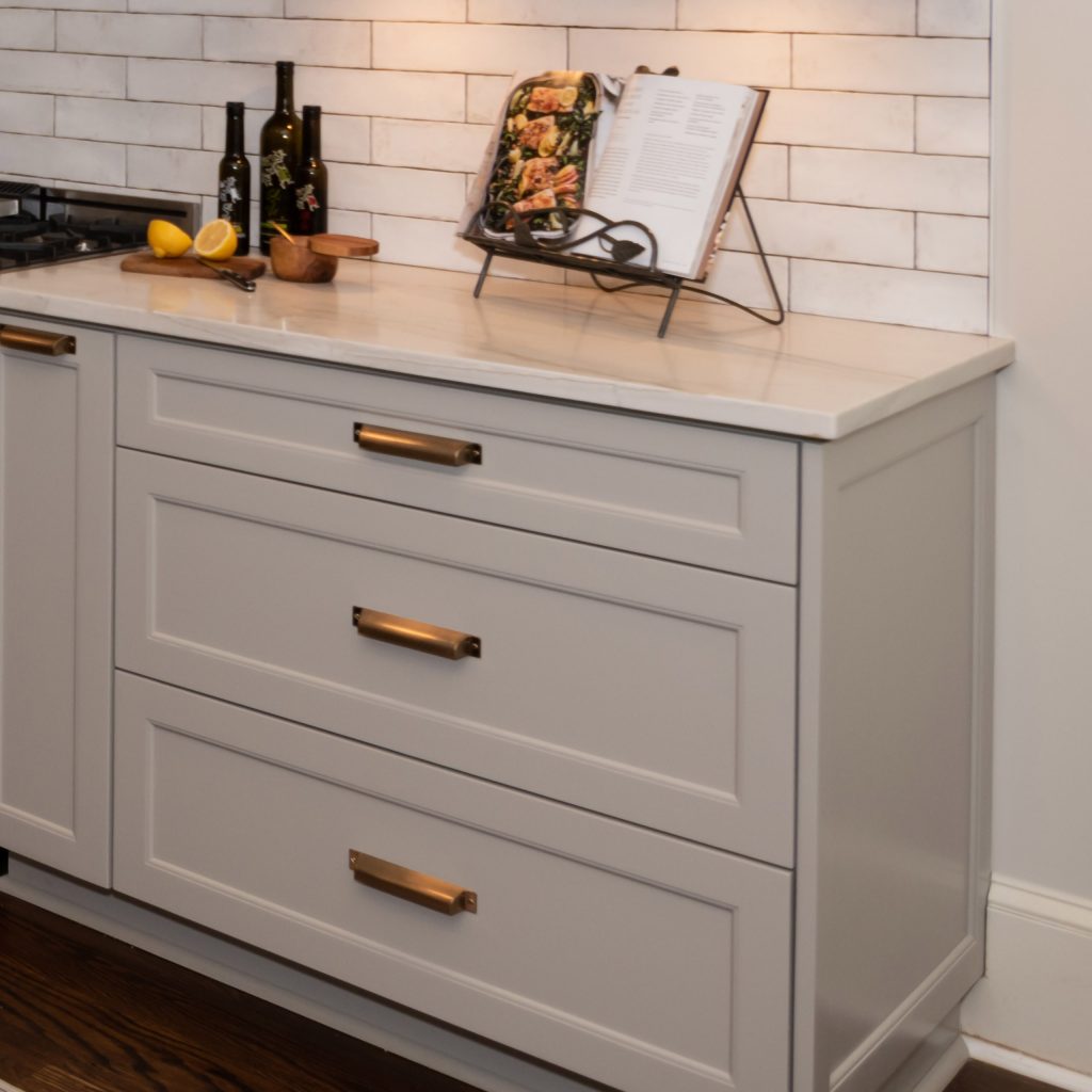 Light-gray drawer base with brass cup pulls, a white quartz top, and handmade white subway tile behind a cookbook stand.