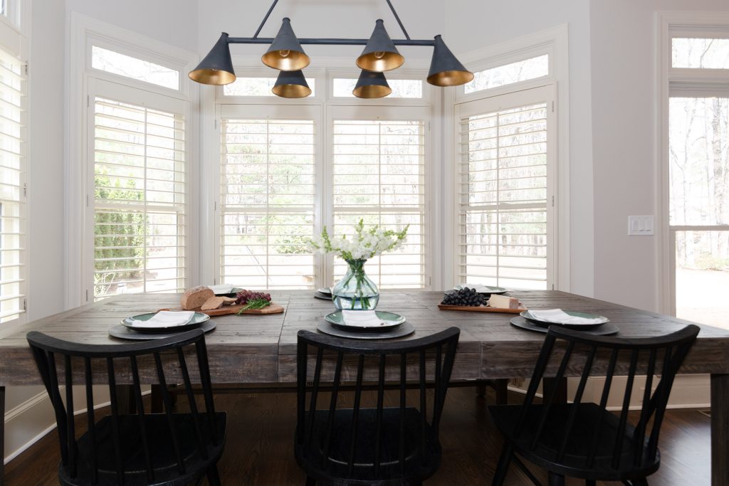 Breakfast nook with a reclaimed-wood table, black spindle chairs, and a black-and-brass chandelier framed by shuttered windows.