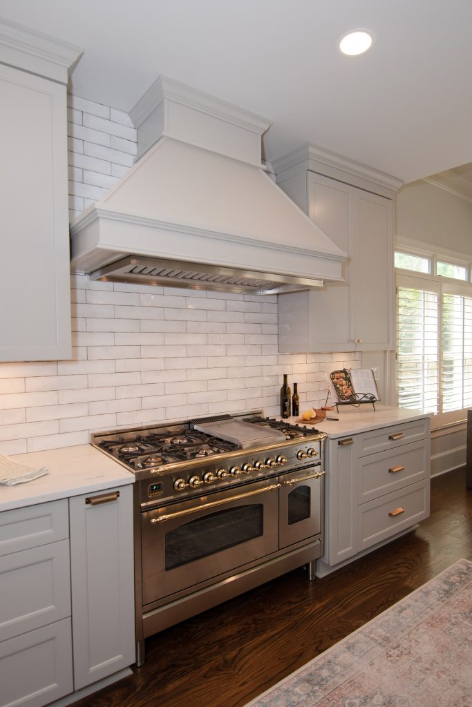Stainless pro range with brass knobs and a painted hood over handmade white subway tile, flanked by light-gray cabinets.