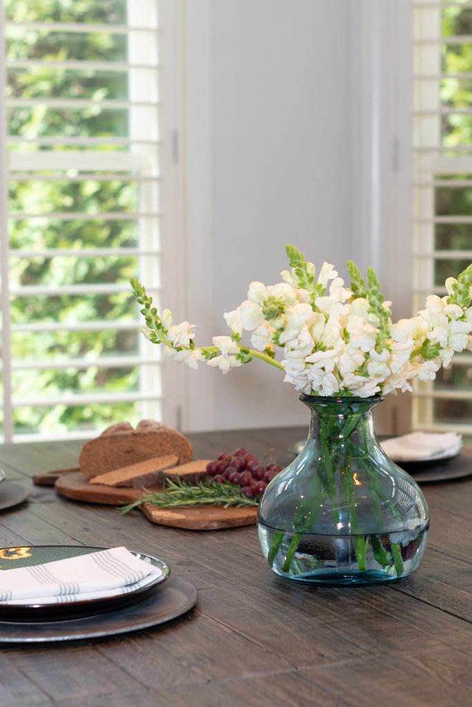 Blue-green glass vase of white snapdragons on a dark reclaimed-wood table with a bread board and red grapes nearby.