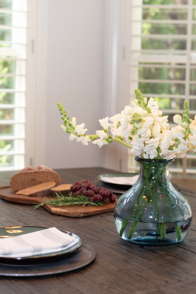 Reclaimed-wood dining table set with pewter chargers, dark plates, striped napkins, and a glass vase of white snapdragons.