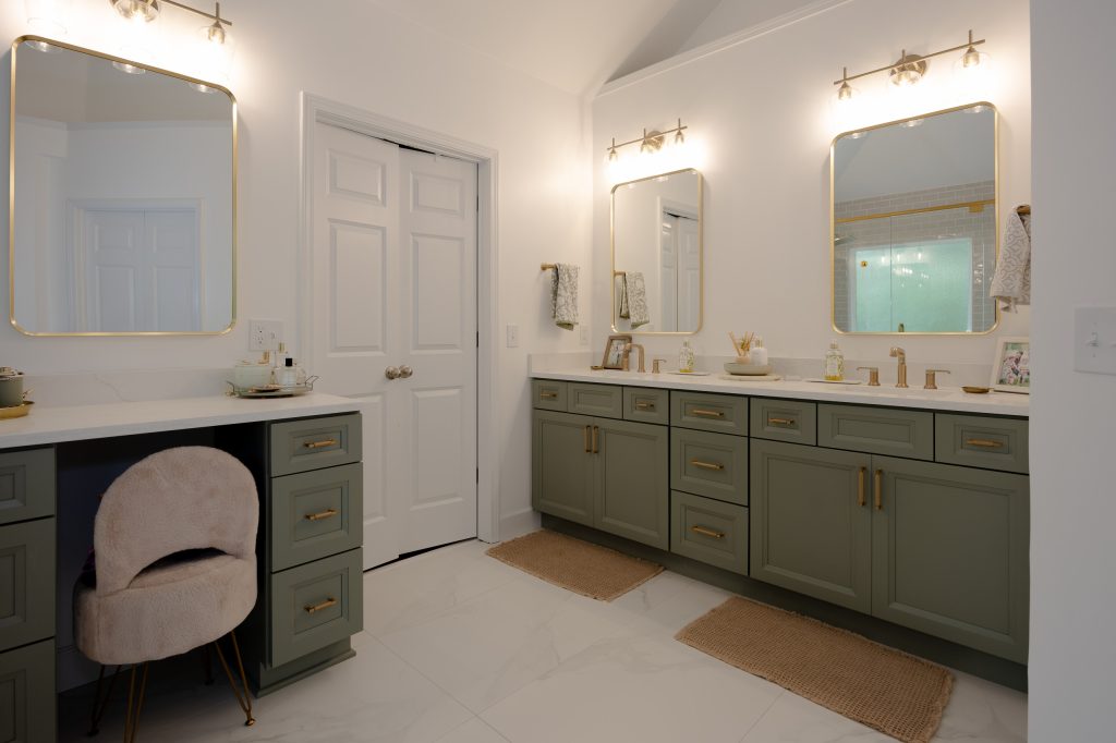 Full master bath view with sage-green vanities, a makeup nook with plush chair, gold mirrors, and a glass shower.