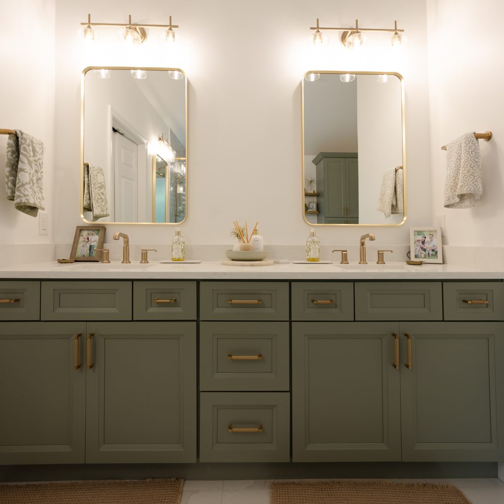 Double vanity in sage-green shaker cabinets with brushed-gold pulls, gold mirrors, and glass-globe sconces.