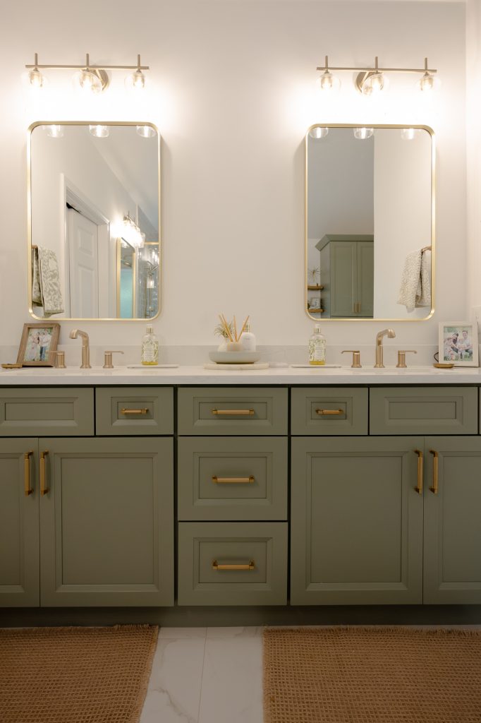 Illuminated double vanity in sage-green shaker cabinets with gold hardware, two gold mirrors, and glass-globe sconces.