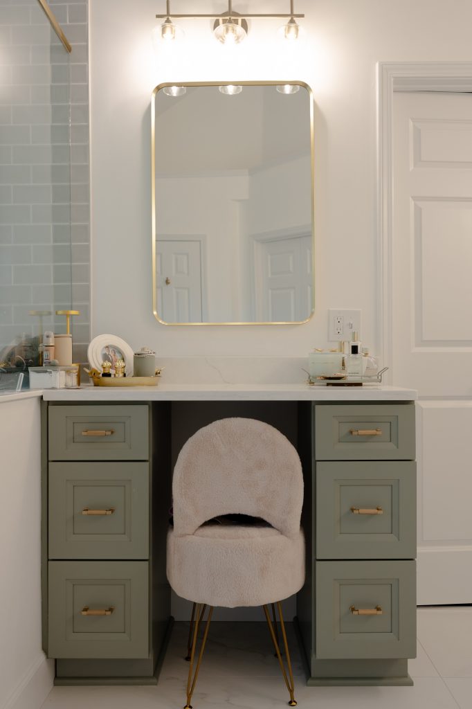 Sage-green makeup vanity with a gold-framed mirror, glass sconce, quartz top, and a plush cream chair on gold legs.