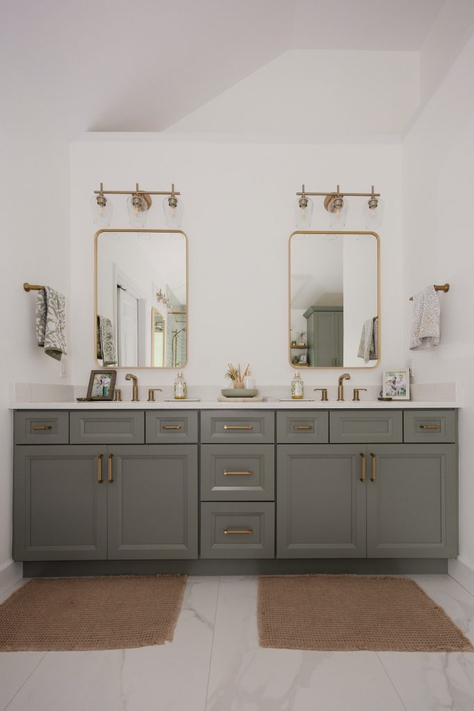 Double vanity in gray-green shaker cabinets with two gold mirrors, gold sconces, quartz counters, and jute rugs.
