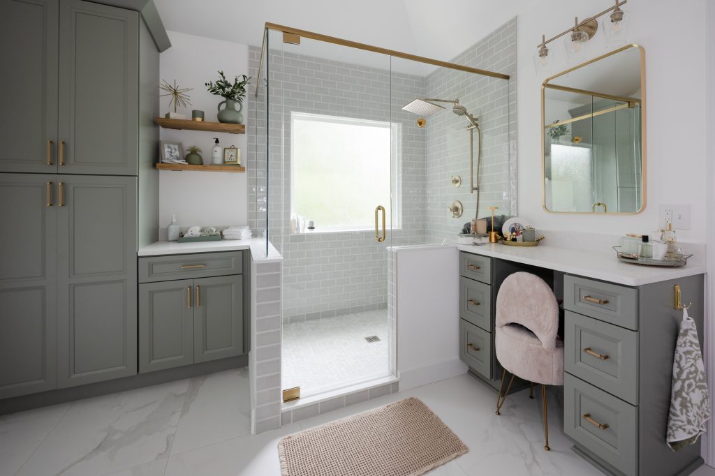 Sage-green master bath with a glass shower, gold sconces and mirror, floating shelves, and a blush makeup-vanity chair.