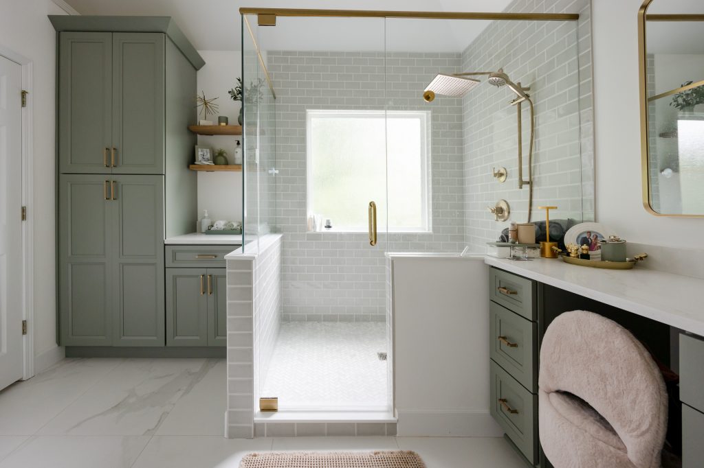 Master bath with sage-green cabinets, frameless glass shower in gray subway tile, and a makeup vanity with a plush chair.