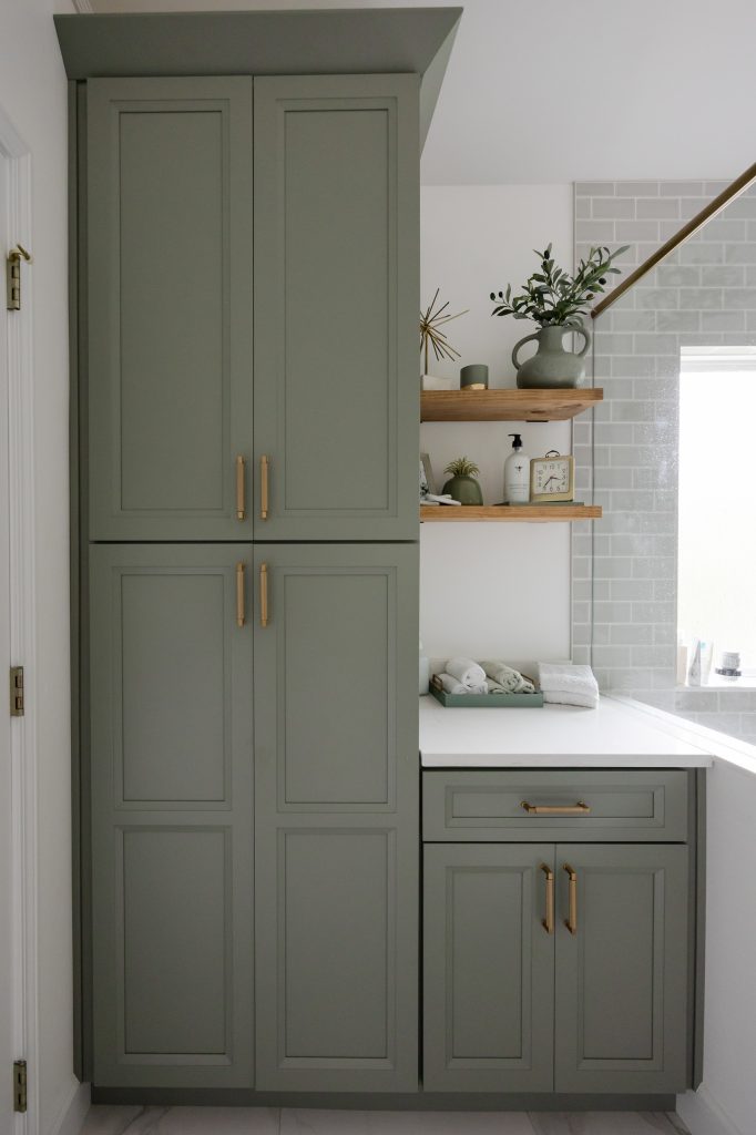 Tall sage-green linen cabinet and lower vanity with brushed-gold hardware, floating wood shelves, and white quartz top.