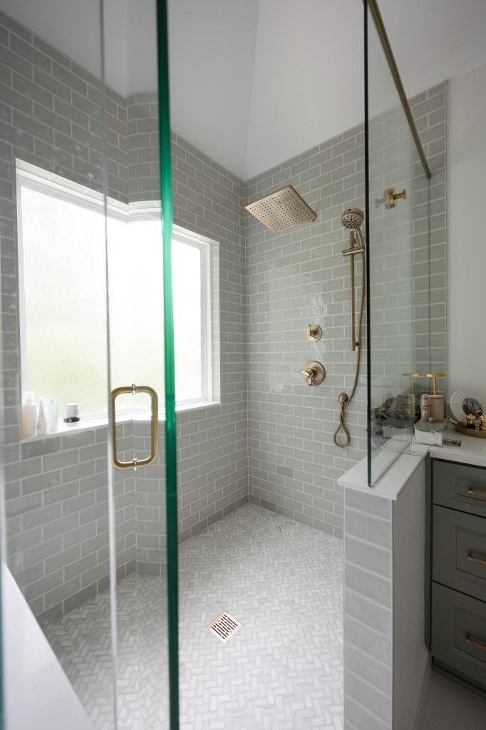 Walk-in shower in gray subway tile with champagne-bronze rain and handheld heads and a marble-look herringbone floor.