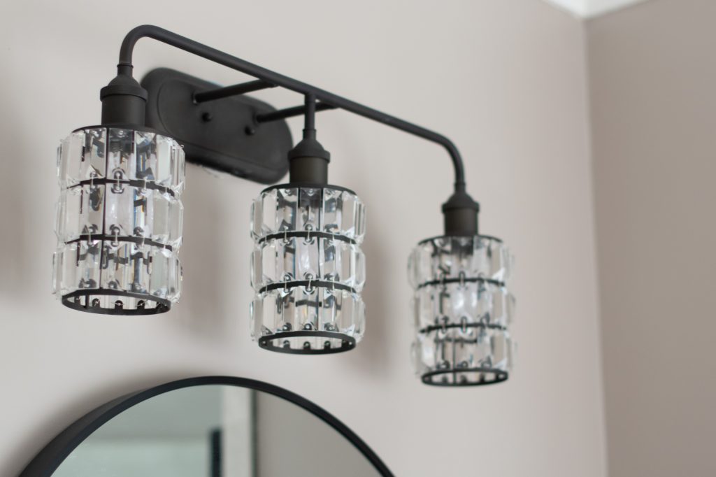 Oil-rubbed bronze three-light vanity fixture with crystal cylinder shades mounted above a round black-framed mirror.
