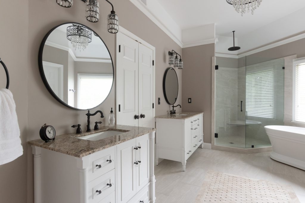 Primary bath with two white furniture-style vanities, speckled quartz tops, round black mirrors, a corner glass shower, and a freestanding tub.