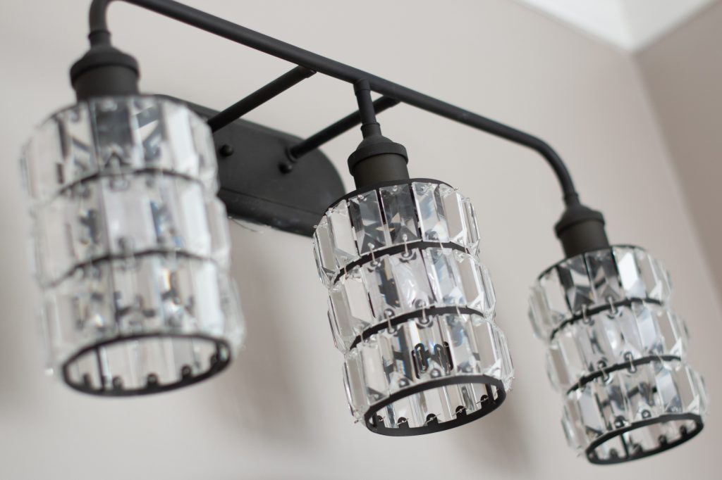 Close-up of an oil-rubbed bronze three-light vanity fixture with faceted crystal cylinder shades against a soft greige wall.