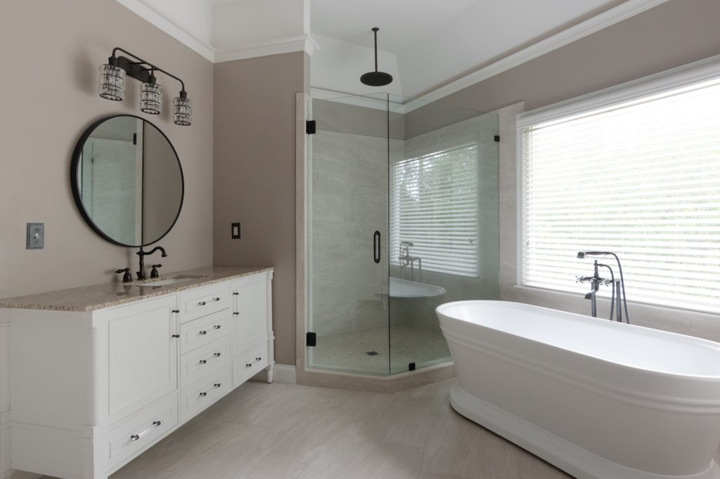 Primary bath with a white vanity, round black mirror, neo-angle glass shower, and a freestanding tub beneath the window.