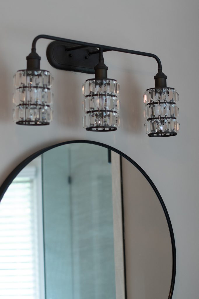 Oil-rubbed bronze three-light vanity fixture with crystal cylinder shades above a round black-framed mirror.