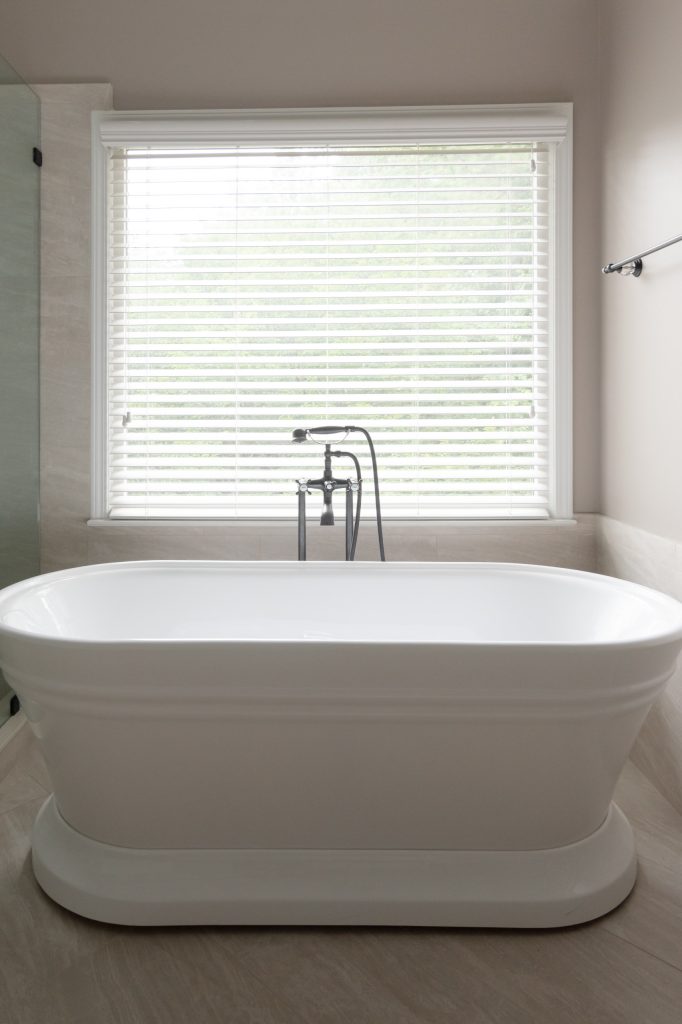 White freestanding double-ended soaking tub centered under a shuttered window with a floor-mounted telephone tub filler.