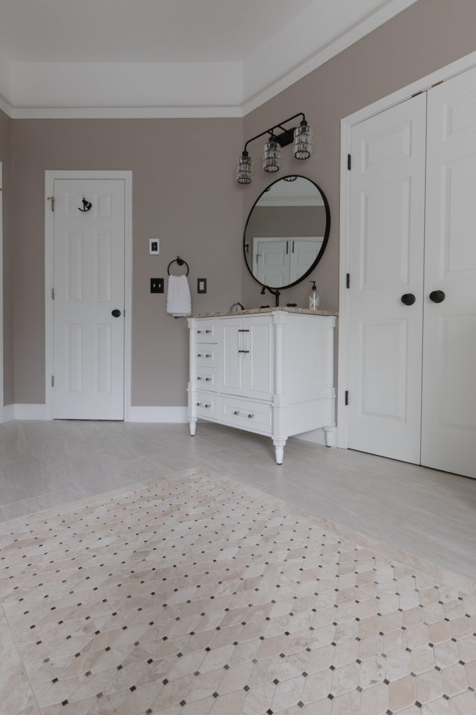 Single white furniture-style vanity with a round black mirror and crystal sconce against a greige wall.