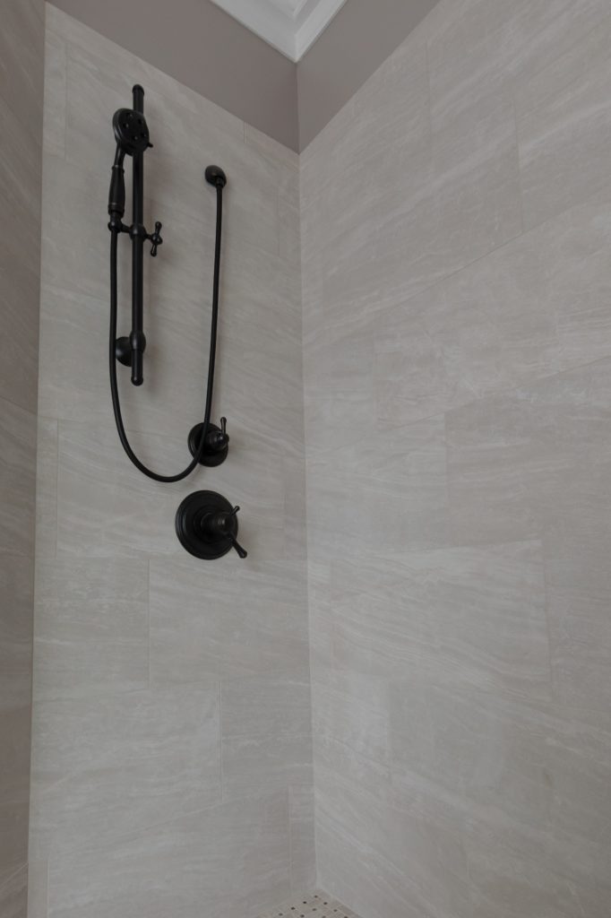 Oil-rubbed bronze slide-bar hand shower and thermostatic valve mounted on pale marble-look shower tile.