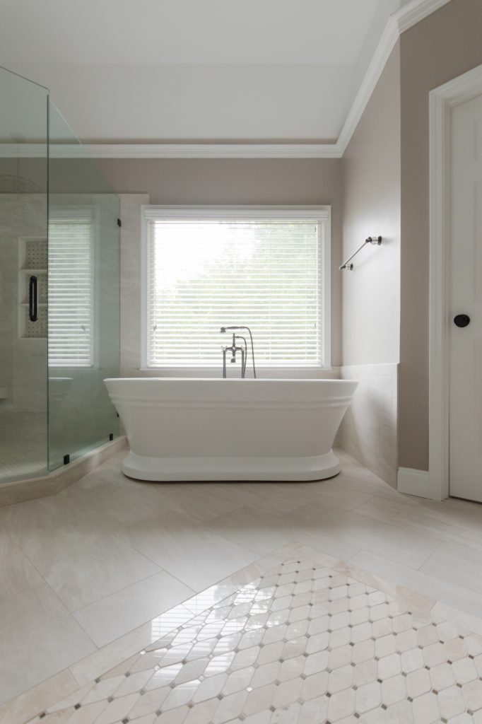 Freestanding white tub beneath a window with an oil-rubbed bronze filler, beside a glass shower over marble-look tile.