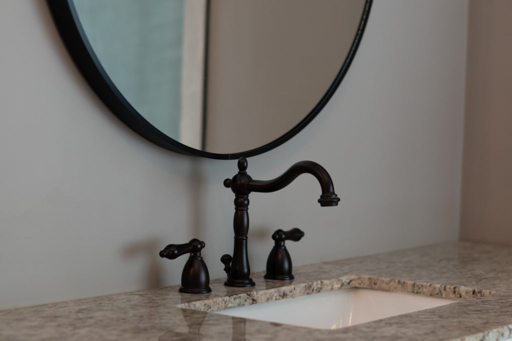 Oil-rubbed bronze widespread faucet with lever handles on a speckled quartz vanity below a round black mirror.