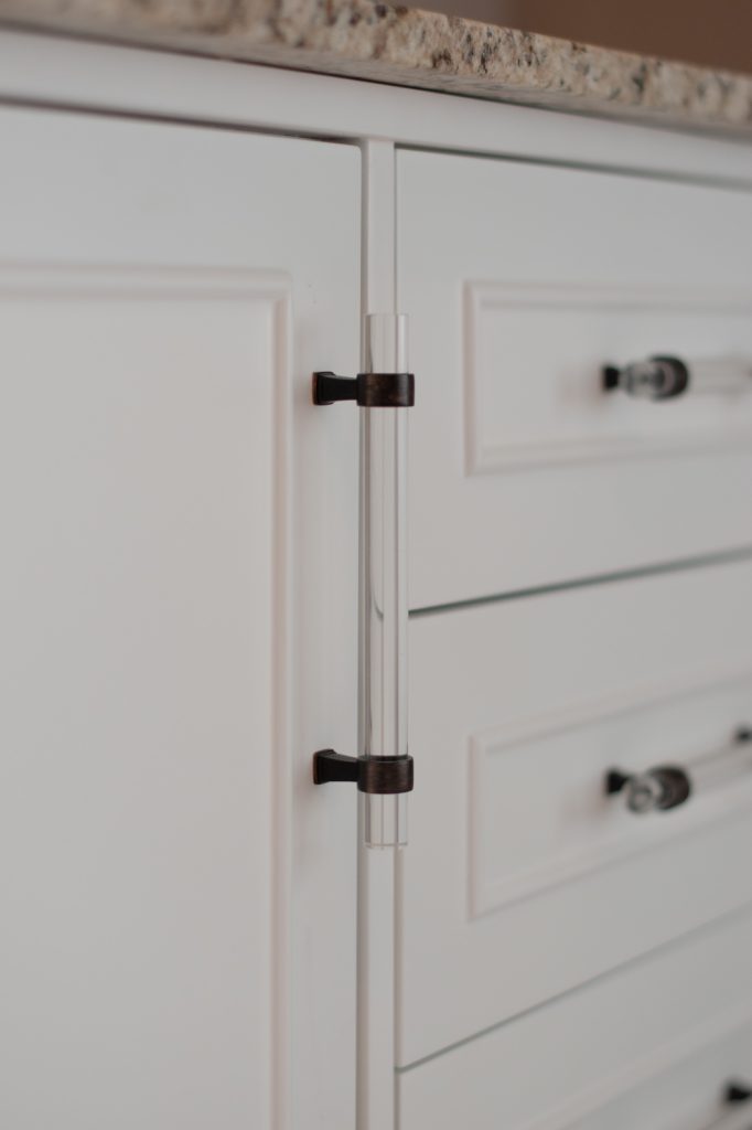 White vanity cabinetry with clear acrylic bar pulls on oil-rubbed bronze mounts under a speckled quartz top.