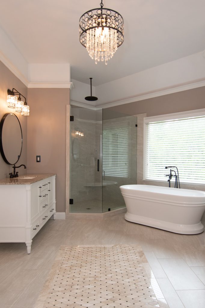 Vertical shot of a luxury bath with a white vanity, crystal drum chandelier, corner glass shower, and freestanding tub.
