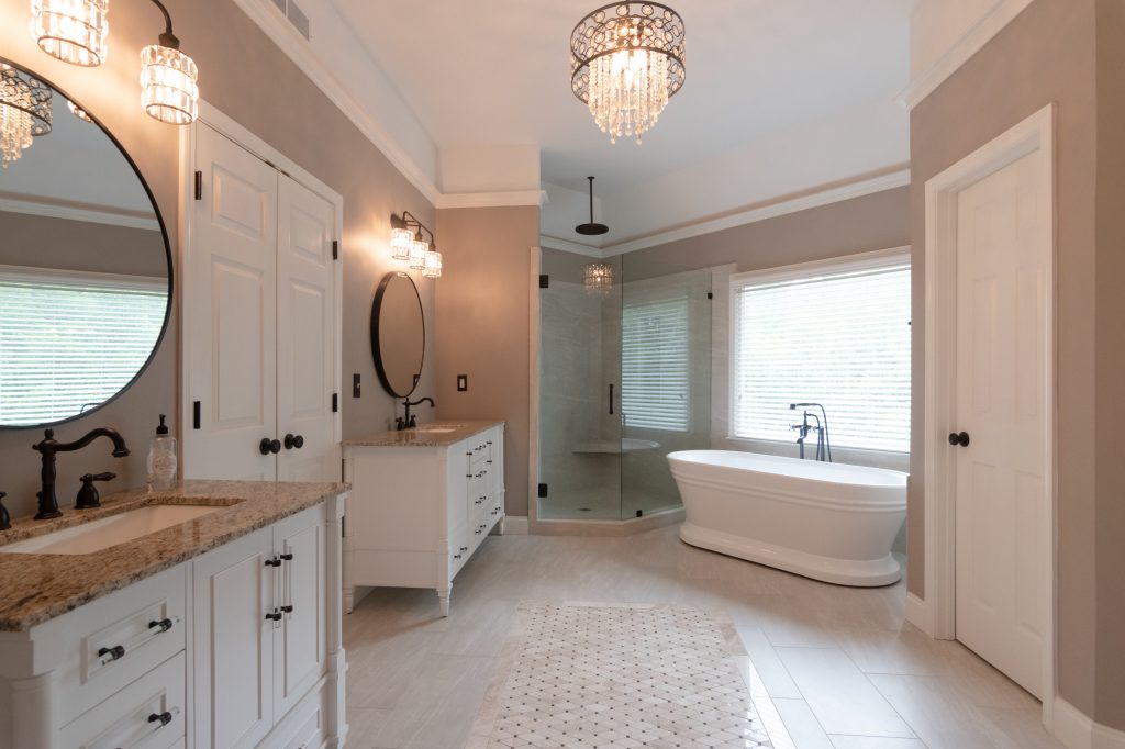 Primary bathroom with twin white vanities, oil-rubbed bronze fixtures, a corner shower, freestanding tub, and crystal chandelier.