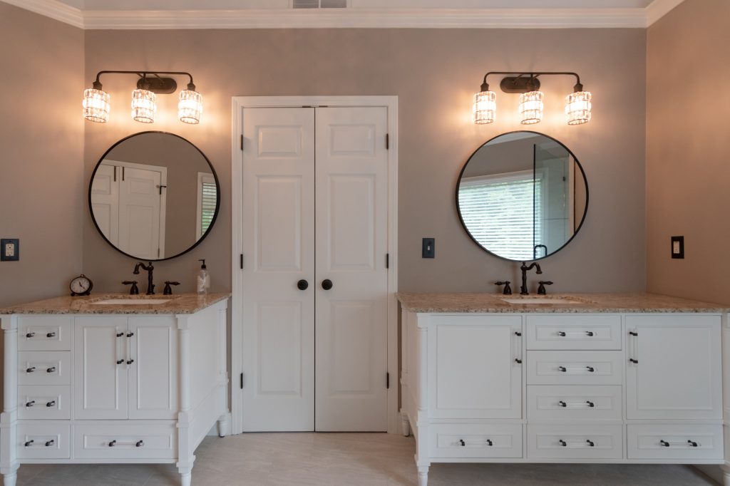Matching pair of white bath vanities with round mirrors and crystal cylinder sconces on either side of a white door.