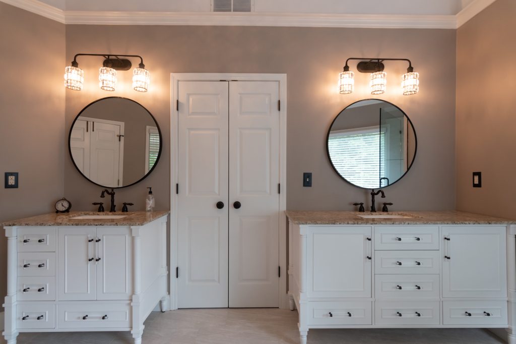 Two matching white vanities with speckled quartz tops, round black mirrors, and crystal sconces flanking a white double door.