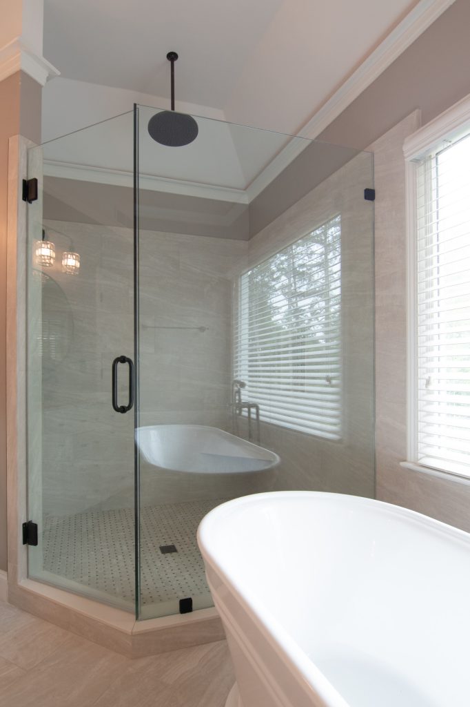 Frameless corner glass shower with a black ceiling-mount rain head, marble-look tile, and a freestanding tub in the foreground.