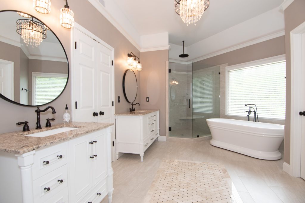 Primary bath showing twin white vanities with speckled quartz tops, round mirrors, a corner shower, and a freestanding tub.