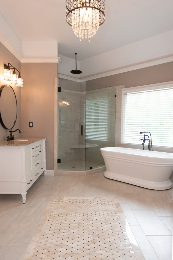 Vertical view of a luxury primary bath with a white vanity, corner glass shower, freestanding tub, and crystal chandelier.