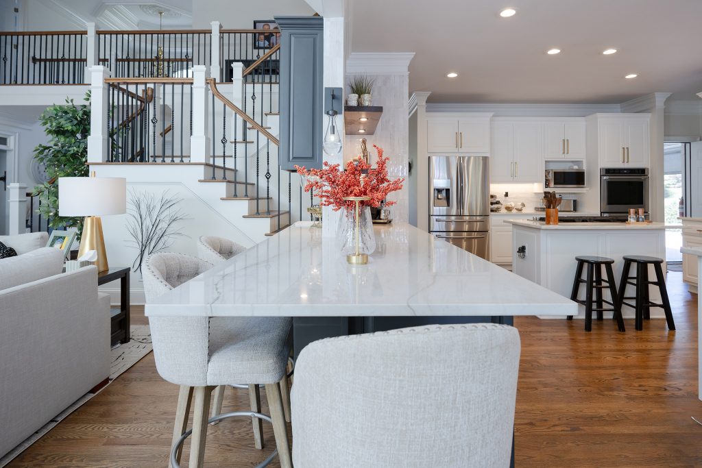 Kitchen island with veined quartz top and gray barstools set against a two-story staircase and white kitchen beyond.