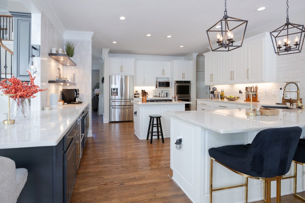 White-and-navy kitchen with brushed-gold pulls, quartz island, black barstools, subway tile, and cage lantern lighting.