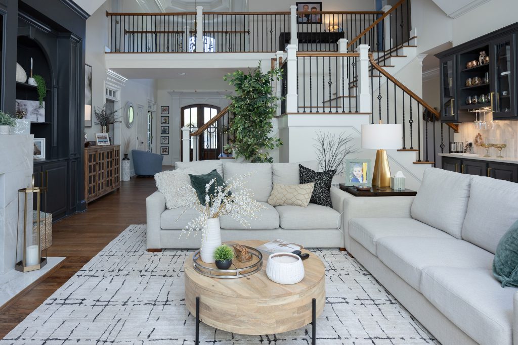 Great room and foyer view with curved staircase, gray sectionals, tall greenery, and a charcoal wet bar.