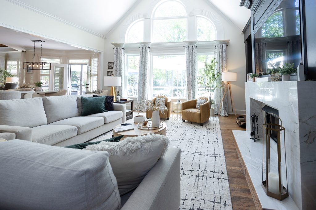Vaulted great room with two-story arched windows, linen sectionals, plantation shutters, and a marble fireplace.